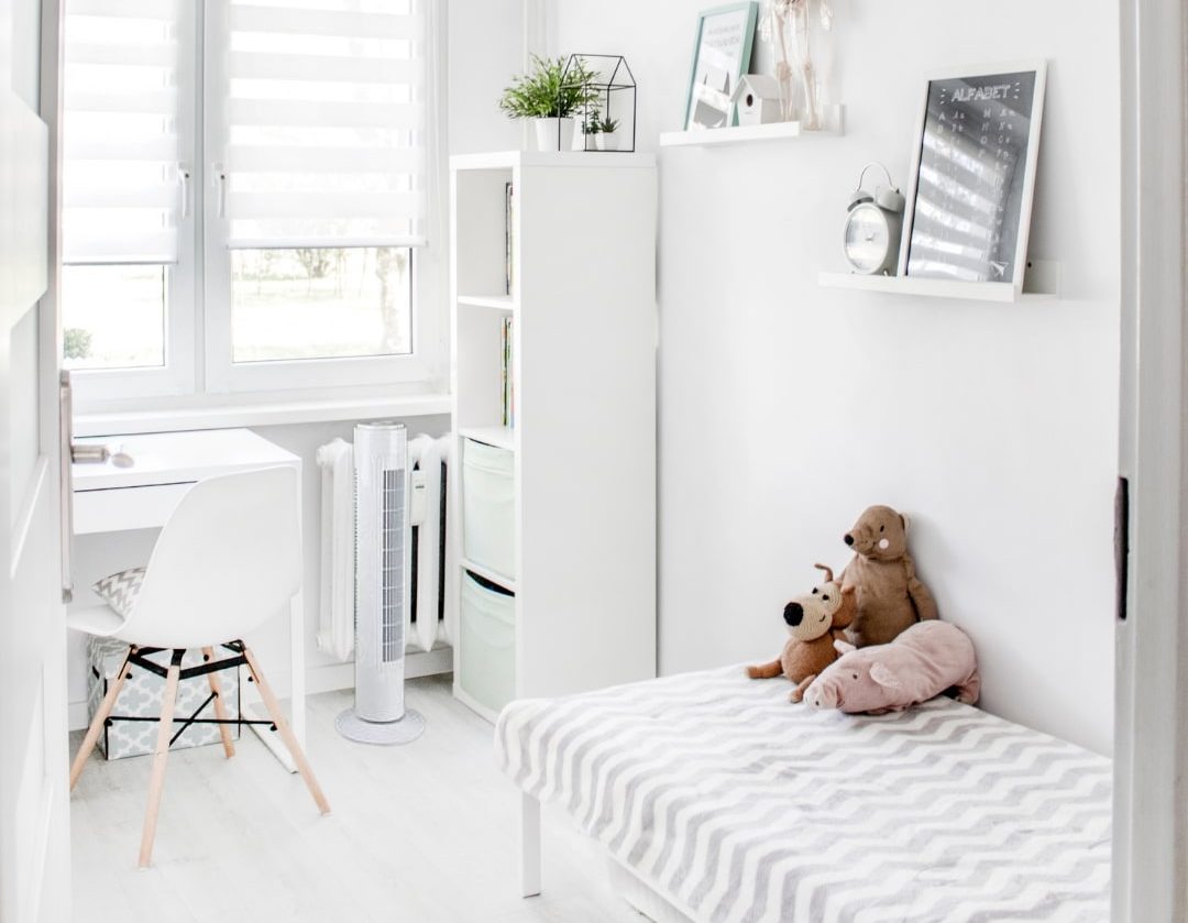 Salle blanche avec un ventilateur sur pied près d’un bureau et d’un lit, dans une pièce lumineuse et décorée avec des