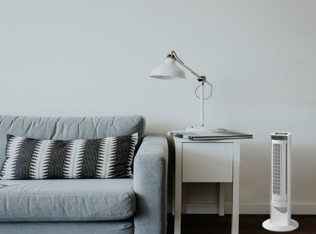 Ventilateur colonne blanc à côté d’une table de chevet et d’un canapé gris dans un séjour.