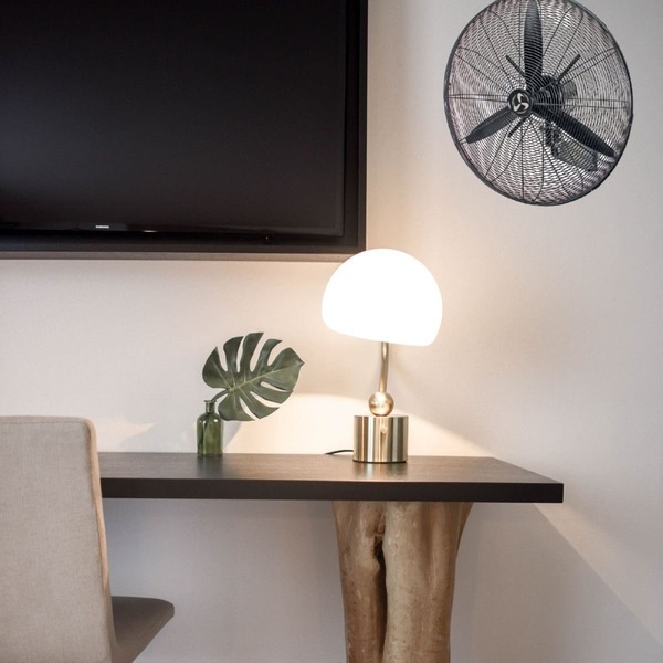 Lampe de table blanche sur un bureau avec ventilateur mural en arrière-plan.