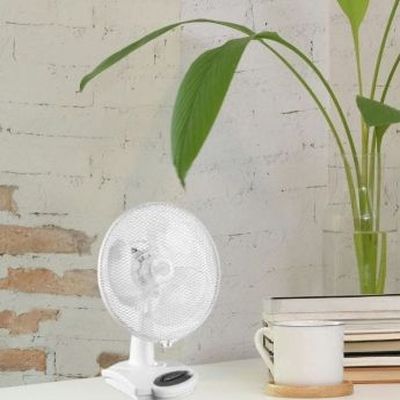 Petit ventilateur rechargeable blanc posé sur une table près d’un mug et de quelques livres, avec une plante en arrière-plan.