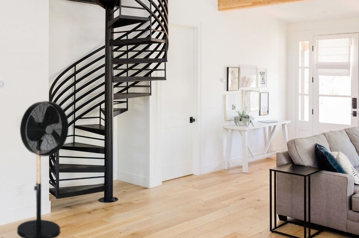 Salon lumineux avec canapé et table basse près d’un escalier en spirale noir, et un ventilateur debout sur le parquet.