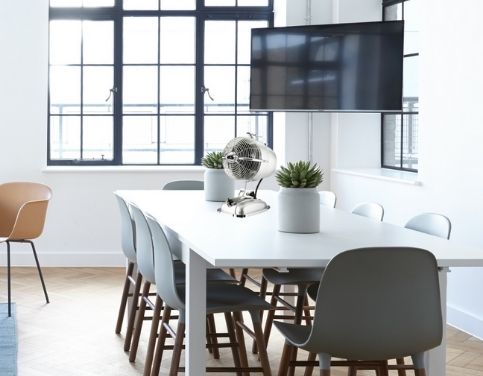 Table blanche entourée de chaises dans une salle lumineuse avec un ventilateur et une télévision au mur.