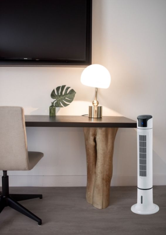 Ventilateur sur pied blanc placé dans un intérieur près d’un bureau avec lampe et plante décorative.
